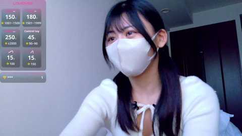 Video of an East Asian woman with long black hair, wearing a white mask, white top, and a pink background, displaying a digital thermometer with readings.