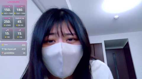 A video of a young Asian woman with long black hair wearing a white mask indoors. The background features a white wall and a ceiling light. A digital screen on the left displays temperature and humidity readings.