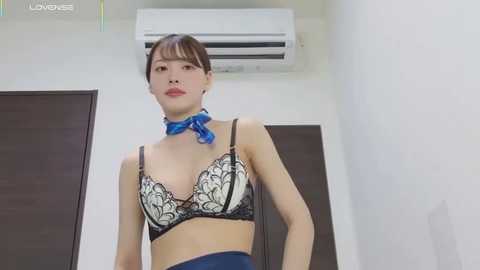 Media: Video of an Asian woman with light skin, short brown hair, and bangs. She wears a black lace bra with blue trim, a blue choker, and blue pants. Background features a white wall with an air conditioner and closed door.