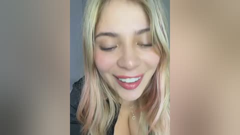 Video of a smiling young woman with fair skin, long blonde hair, and a small pendant necklace. She wears a black top, with a blurred background.