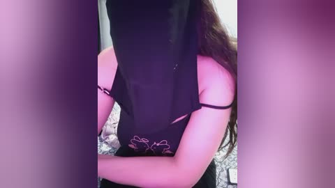 Video of a woman with long, wavy brown hair, wearing a black lace bodysuit with a floral design, and a black face mask, standing in a bedroom with a patterned bedspread.