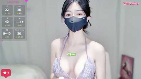 Video of a young Asian woman with pale skin, black hair, and a black mask, wearing a lavender lace bra, in a dimly lit room with a white wall and a pink poster.