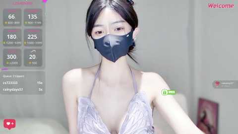 A video of an East Asian woman with fair skin and dark hair, wearing a black face mask and a lacy white top, standing indoors with a social media overlay showing 165 likes and 30 comments.