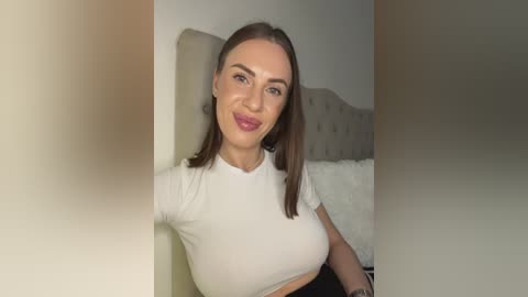 Video of a light-skinned woman with shoulder-length brown hair, wearing a white crop top, sitting on a bed with a beige headboard.