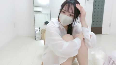 Video of an Asian woman in a white medical gown, sitting on a tiled floor, wearing a surgical mask. Background shows a bathroom with a mirror, toilet, and sink.