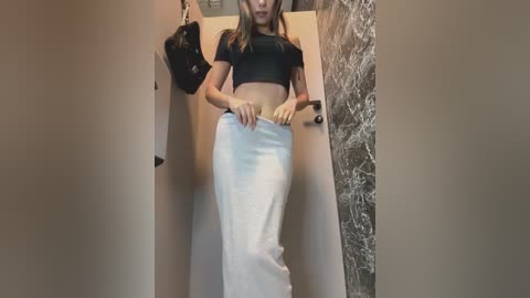 Video of a slender woman with light skin, wearing a black crop top and white high-waisted skirt, standing in a beige hallway with a textured wall and a black handbag on the left.
