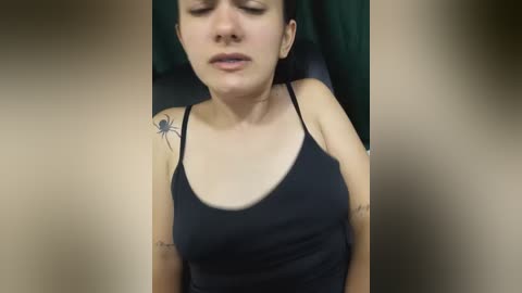 Video of a woman with light skin, dark hair, wearing a black tank top, with a spider tattoo on her left shoulder. She has a relaxed expression.