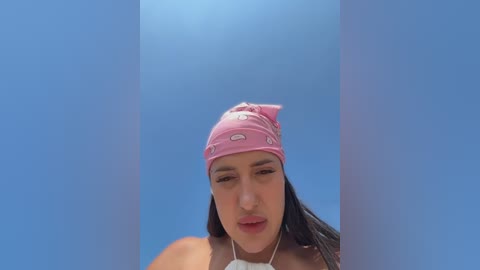 Video of a young woman with olive skin and long dark hair, wearing a pink bandana, white top, and headphones, against a light blue background.