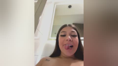 A video of a young Asian woman with long black hair, wearing no top, sticking out her tongue, taken indoors with a white wall and a mirror in the background.
