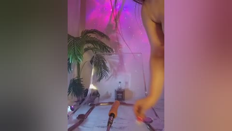 Video of a person in a dimly lit room with purple and pink ambient lighting, holding a long, orange object. A potted palm tree and a bottle are visible in the background.