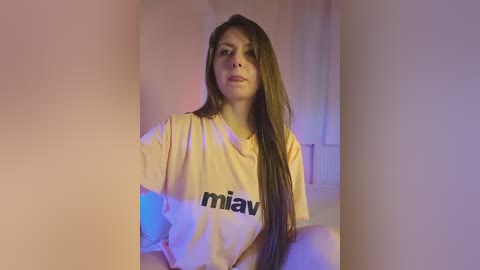 Media: Video of a young woman with long, straight brown hair, wearing a large, light yellow t-shirt with \"miau\" printed in black. She has fair skin and sits in a softly lit room with soft purple and blue hues.
