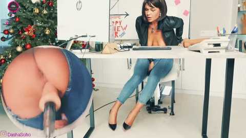 Video of a woman in a black leather jacket, tight jeans, and heels, sitting at a desk in an office with a Christmas tree, a laptop, and a blurred close-up of her crotch.