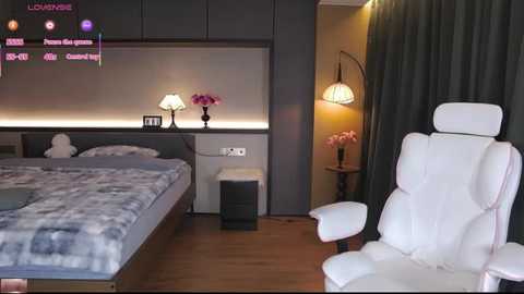 Video of a modern bedroom with a grey headboard, white leather chair, dark curtains, and a cozy bed with a white comforter.
