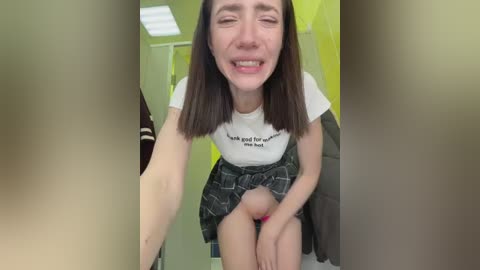 Video of a smiling, fair-skinned woman with shoulder-length brown hair, wearing a white T-shirt with a black slogan and a patterned skirt. She's seated with legs spread, revealing her genital area. Background shows a green-walled room with a coat rack.