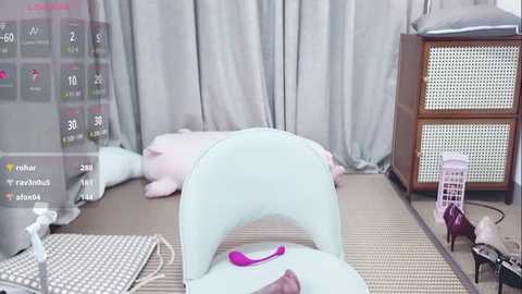 Video of a serene room with a light-blue chair, pink vibrator on a beige mat, grey curtains, wooden cabinet, and a white pillow on the floor.