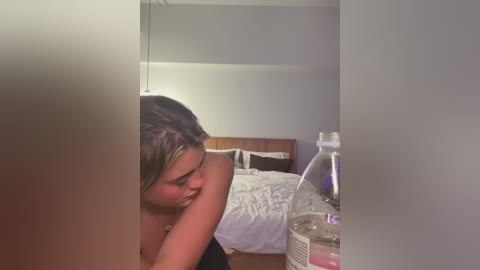 Media: Video of a young woman with medium skin tone and short, dark hair, sitting shirtless in a dimly lit bedroom with a wooden headboard, bed, and a large water bottle.