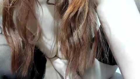 A close-up video of a woman's chest, showing her light skin and long, wavy brown hair partially covering her breasts. The background is out of focus, emphasizing the textures of her hair and skin.