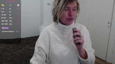 Video of a woman with shoulder-length blonde hair, wearing a white textured sweater, holding a phone, in a minimalistic bedroom with a dark bedspread.