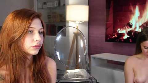 Video of a topless woman with long red hair and a tattoo, standing next to a transparent chair in a modern living room with a lit fireplace and white shelves in the background.