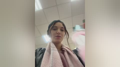 A video of a young woman with light skin and dark hair, wearing a black jacket and a pink scarf, standing in a bathroom with a white ceiling and fluorescent lights.