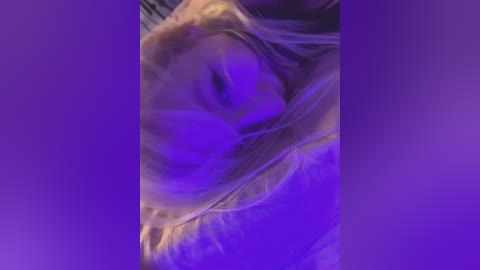 A close-up video with a purple hue filters a person's face, featuring long, blonde hair and a partially visible nose and mouth. The background is blurred, with a gradient from light to dark purple, enhancing the ethereal and intimate atmosphere.