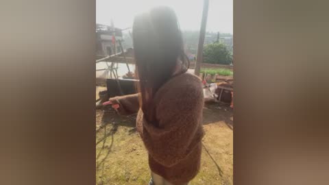 Video of a woman with long black hair, wearing a brown sweater, holding a pink phone, standing outdoors near a construction site with a blurry background.