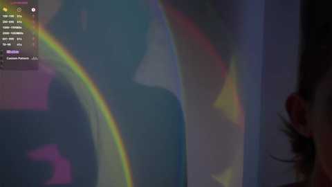 A video captures a person's silhouette with a vibrant rainbow projected behind them. The figure appears to be seated, and the setting has a soft, ambient light with a blurred background.