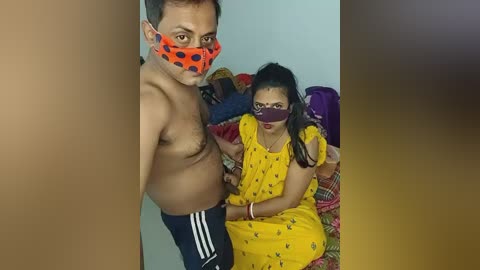 Video of a shirtless man with a polka-dot mask and a woman in a yellow sari, both with masks, sitting on a bed in a dimly lit room.