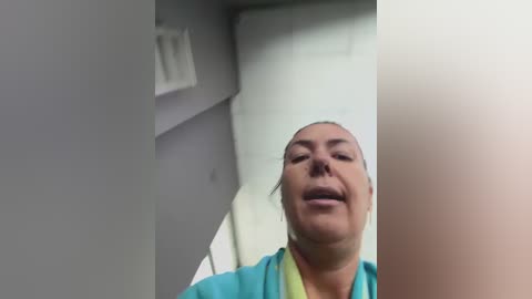 A video of a middle-aged, light-skinned woman with a fair complexion, wearing a turquoise shirt, smiling with closed eyes. The background shows a white wall and a partially visible white door.