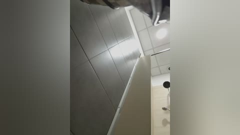 A video of a narrow, white-tiled bathroom ceiling and wall, featuring a round, white ceiling light fixture and a corner shower stall. The space appears clean and utilitarian.