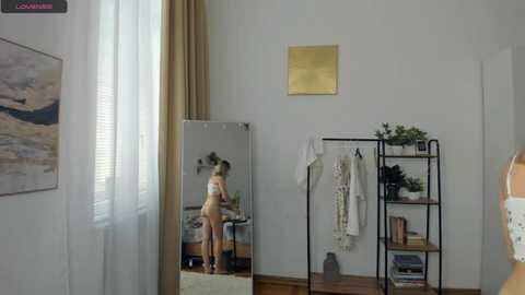 Video of a minimalist bedroom with a nude woman in front of a mirror, beige curtains, white walls, and modern decor.