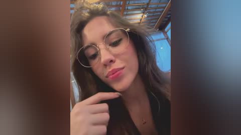 A video of a young woman with long brown hair and glasses, puckering her lips, sitting indoors with wooden beams and blue sky visible.