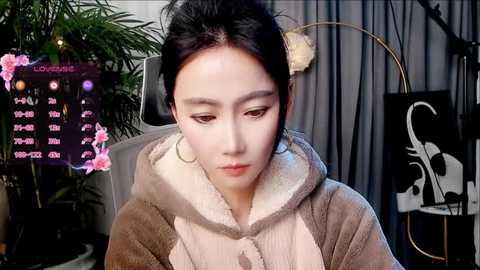 A video of an East Asian woman with pale skin, dark hair, and expressive eyes, wearing a brown fur-lined hoodie, seated indoors. The background features a potted plant, a computer screen displaying weather info, and a wall with a black and white graphic.