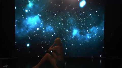 A video of a nude woman with blonde hair, sitting on the floor, gazing at a large, projected image of the Milky Way galaxy, creating a serene, introspective atmosphere.