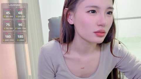 A video of an East Asian woman with straight, shoulder-length brown hair, wearing a light grey top. She has a neutral expression. Background includes a window and a chair. A \"LoverLive\" overlay shows temperature and heart rate.