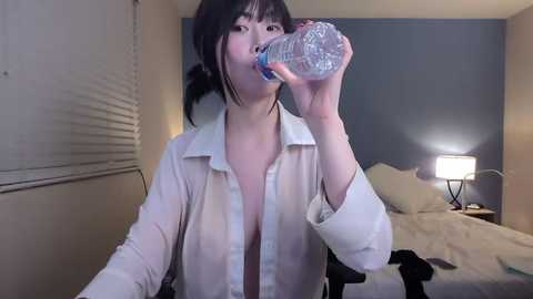 Video of an Asian woman with short black hair, wearing a see-through white blouse, drinking from a plastic bottle in a dimly lit bedroom with a bed, lamp, and closed blinds.