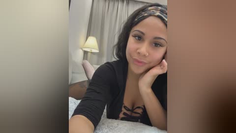 Video of a young Latina woman with medium skin tone, long black hair, and a nose ring, lying on a bed with a black bra, black cardigan, and a colorful headband. The room features a lamp and curtains.