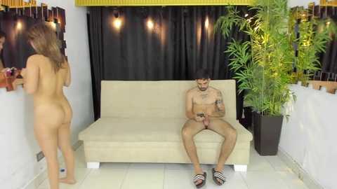 Video: Nude man with tattoos on sofa, shirtless woman in sandals and braids, dark curtains, bamboo plant, wooden shelves, indoor setting.