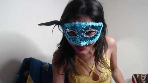 Video of a woman with long dark hair wearing a blue feathered masquerade mask, yellow lace top, and red lipstick, against a plain white wall background.