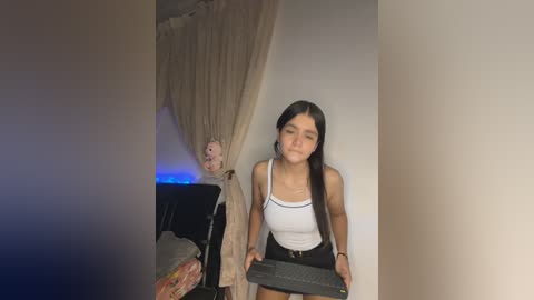 Video of a young woman with long black hair, wearing a white tank top and black shorts, standing in a dimly lit room with a bed and a curtain.