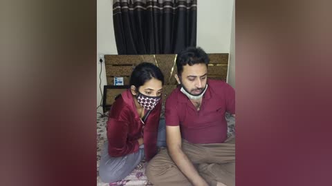 Media: Video of a young South Asian couple in a bedroom; man with short dark hair and beard, wearing a maroon polo, beige pants, and a face mask; woman with long dark hair, maroon jacket, and face mask, sitting on a bed with patterned bedding.