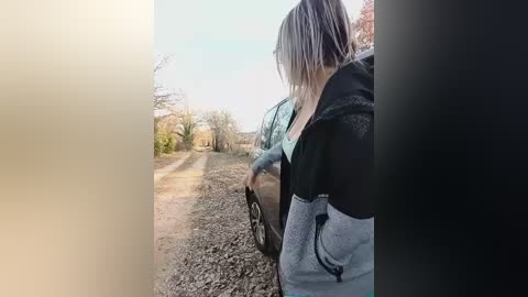 A video of a woman with shoulder-length, platinum blonde hair, wearing a black and grey hoodie, standing beside a parked car on a dirt path in a rural setting.