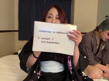 Video of a young Asian woman with long, dark hair, wearing a black robe, holding a sign reading \"Kabukichan_in/aobabozu\" and \"X-amount! @kabukichan\" in a hotel room with beige walls and dark curtains.