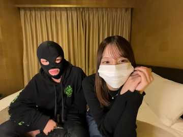 Video of two Asian women, one in a black hoodie with a mask, the other in a black mask and a white mask, sitting on a beige bed, with beige curtains in the background.