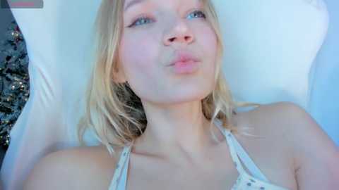 A video of a young blonde woman with fair skin, blue eyes, and a small nose, puckering her lips in a kiss. She is wearing a white, patterned spaghetti-strap top. The background is a white pillow and a partially visible patterned blanket.
