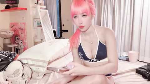 Video of a young woman with pink hair in a dark blue bikini, sitting on a bed in a messy bedroom with posters and pillows.