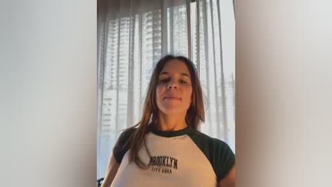 Video of a young woman with long brown hair, wearing a Brooklyn Industries t-shirt, standing in front of a window with sheer white curtains.