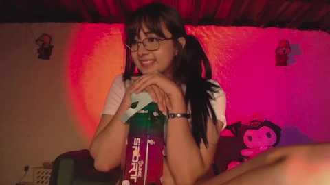 Video of a young Asian woman with long black hair in pigtails, wearing glasses and a white t-shirt, holding a green bottle, against a pink-lit wall with a stuffed toy and phone in the background.
