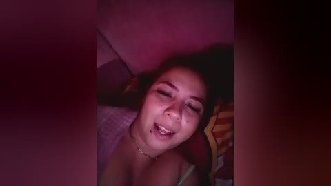 A dimly lit video of an Asian woman with medium skin tone and dark hair, smiling, wearing a colorful striped top, lying on a bed with red and yellow pillows.