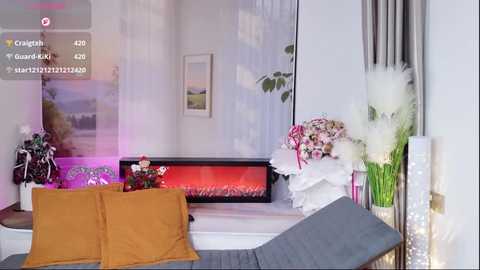 Video of a modern, cozy living room with a fireplace, white sofa, orange and gray throw pillows, floral arrangements, and decorative plants.